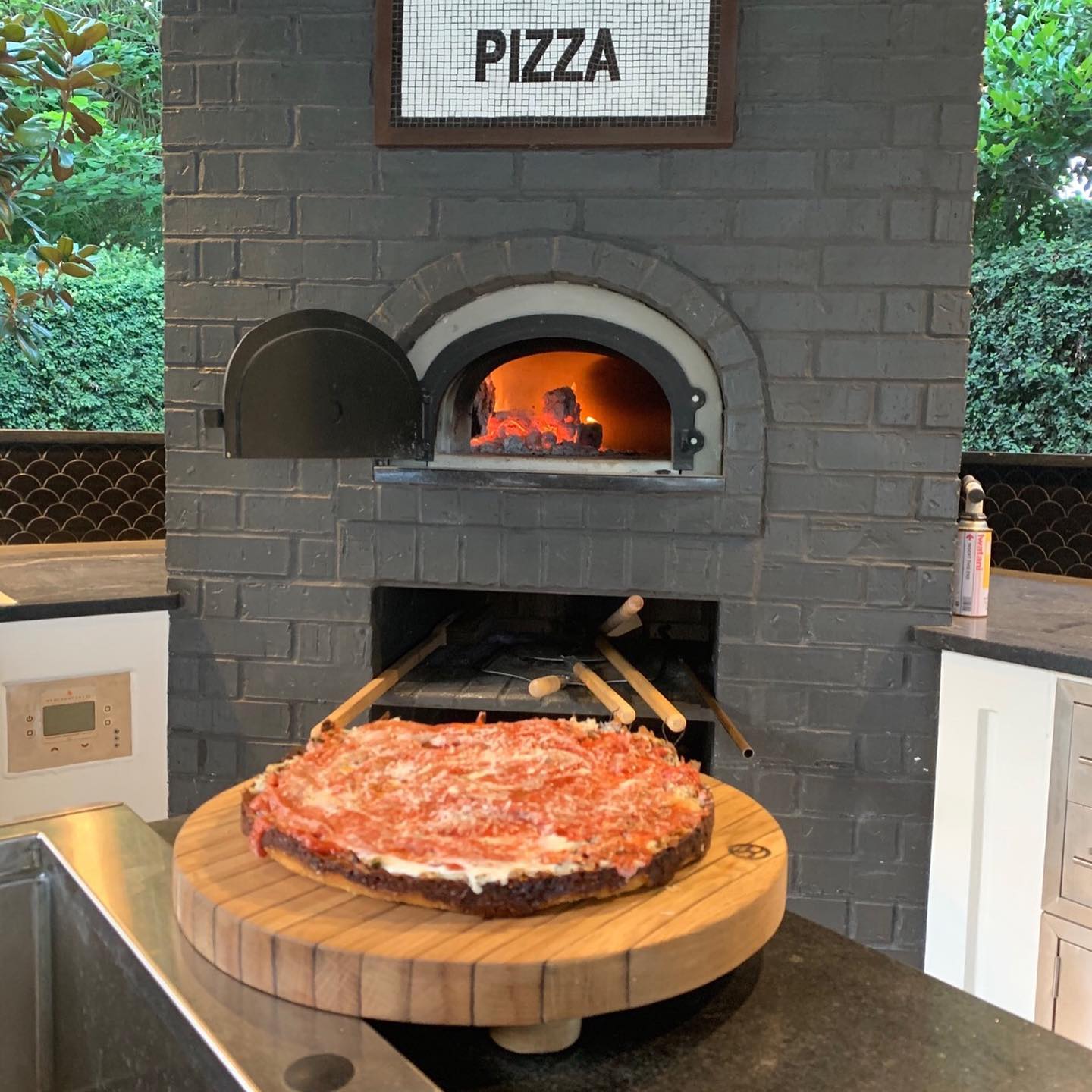 A deep-dish pizza placed on a wooden board in front of a Chicago Brick Oven CBO 750 DIY Kit, our most popular wood-fired pizza oven bundle with a 38" x 28" cooking surface, featuring an intense fire burning inside. A sign labeled "PIZZA" is mounted above the oven. The kitchen setting includes countertops, cabinets, and various cooking utensils.