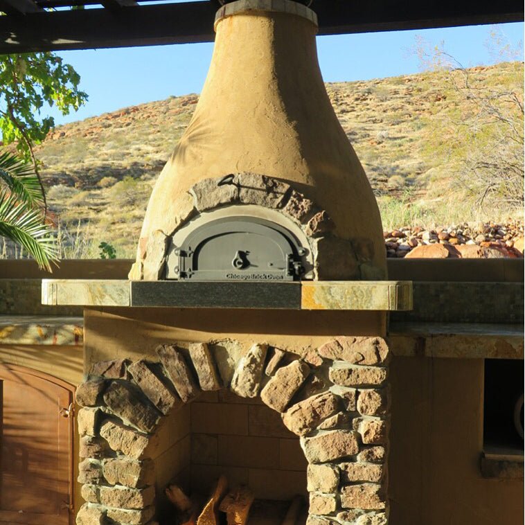 A rustic Chicago Brick Oven CBO 500 DIY kit, made of stone and stucco, sits under a pergola. Firewood is stored in an arch-shaped compartment beneath the wood-fired pizza oven, which features a flexible and affordable 27" x 22" cooking surface. In the background lies a dry, hilly landscape with sparse vegetation.