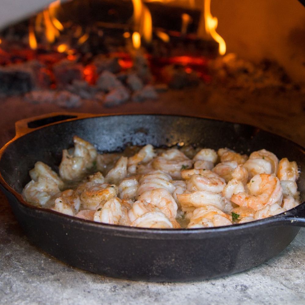 A cast-iron skillet filled with seasoned shrimp is cooking in the high-performance CBO 1000 DIY Kit Wood Fired Pizza Oven by Chicago Brick Oven. The flames and hot coals in the background add a warm, rustic ambiance to the scene. The shrimp are slightly pink, indicating they are nearing readiness on the spacious 53" x 39" cooking surface.