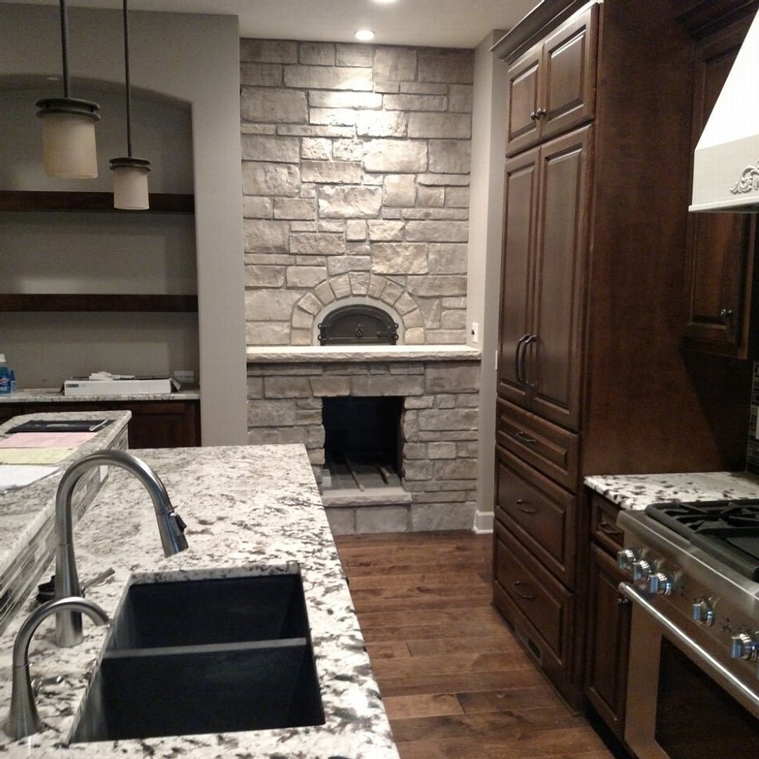 A modern kitchen showcasing a rustic Chicago Brick Oven CBO 500 DIY Kit | Wood Fired Pizza Oven, seamlessly built into a stone wall, reminiscent of the finest outdoor pizza ovens. The granite countertop at the forefront comes with an integrated sink and faucet. Dark wooden cabinets and shelves enhance the stonework and coordinate perfectly with the dark appliances. The room is illuminated by hanging pendant lights, creating a warm ambiance.
