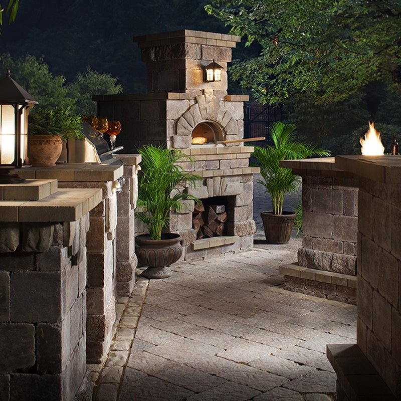 An outdoor kitchen area at dusk showcases the Chicago Brick Oven CBO 1000 DIY Kit wood-fired pizza oven, complete with firewood storage underneath. The setup is surrounded by brick countertops and lush greenery. A fire feature and potted plants enhance the rustic ambiance, adding warmth to the setting.