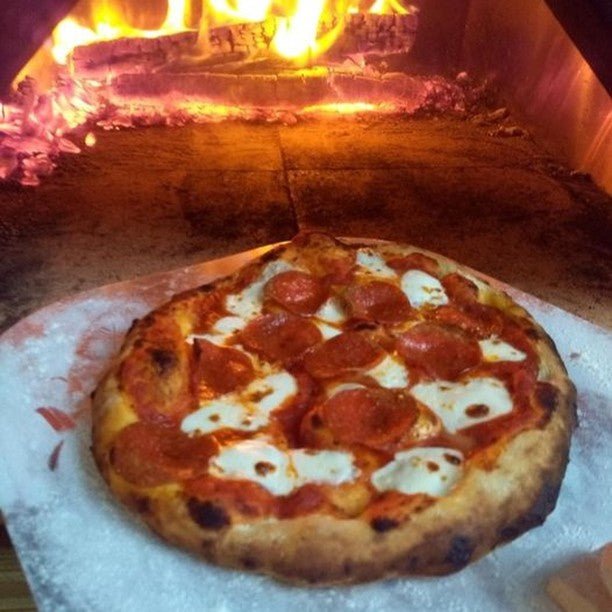 A pepperoni pizza with melted cheese and a slightly charred crust is being baked in the Chicago Brick Oven CBO 750 Tailgater, a mobile wood-burning oven trailer. Flames flicker in the background, enhancing the pizza's rustic and appetizing appearance on its 38" x 28" cooking surface.