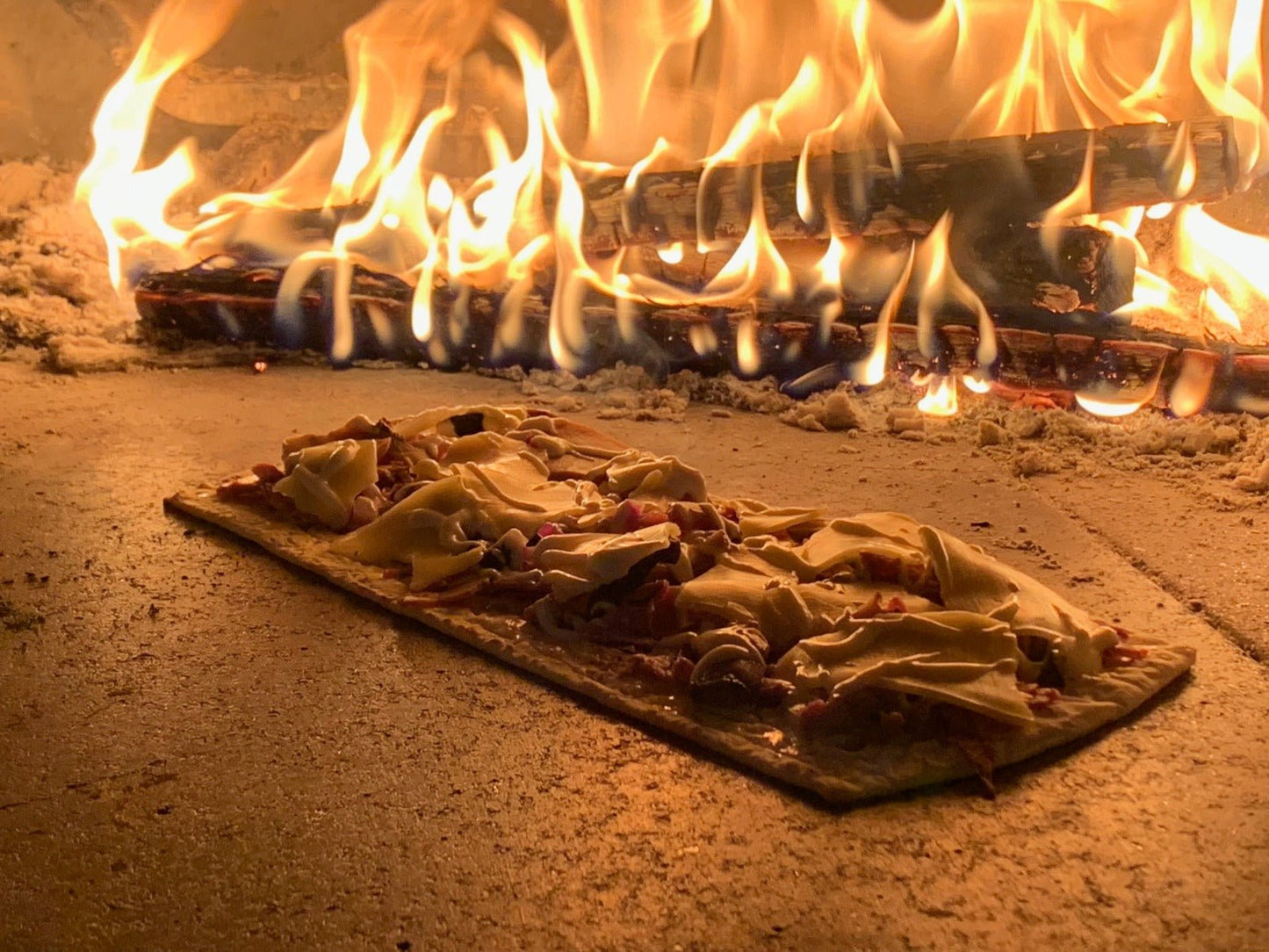 In the Chicago Brick Oven CBO 750 Hybrid DIY Kit (Commercial), a rectangular pizza with melted cheese and toppings bakes on a stone surface in an outdoor pizza oven. Flames and burning wood can be seen at the back, with the oven's intense heat creating visible warmth and a rustic atmosphere. This versatile dual-fuel oven uses both gas and wood for an authentic cooking experience.