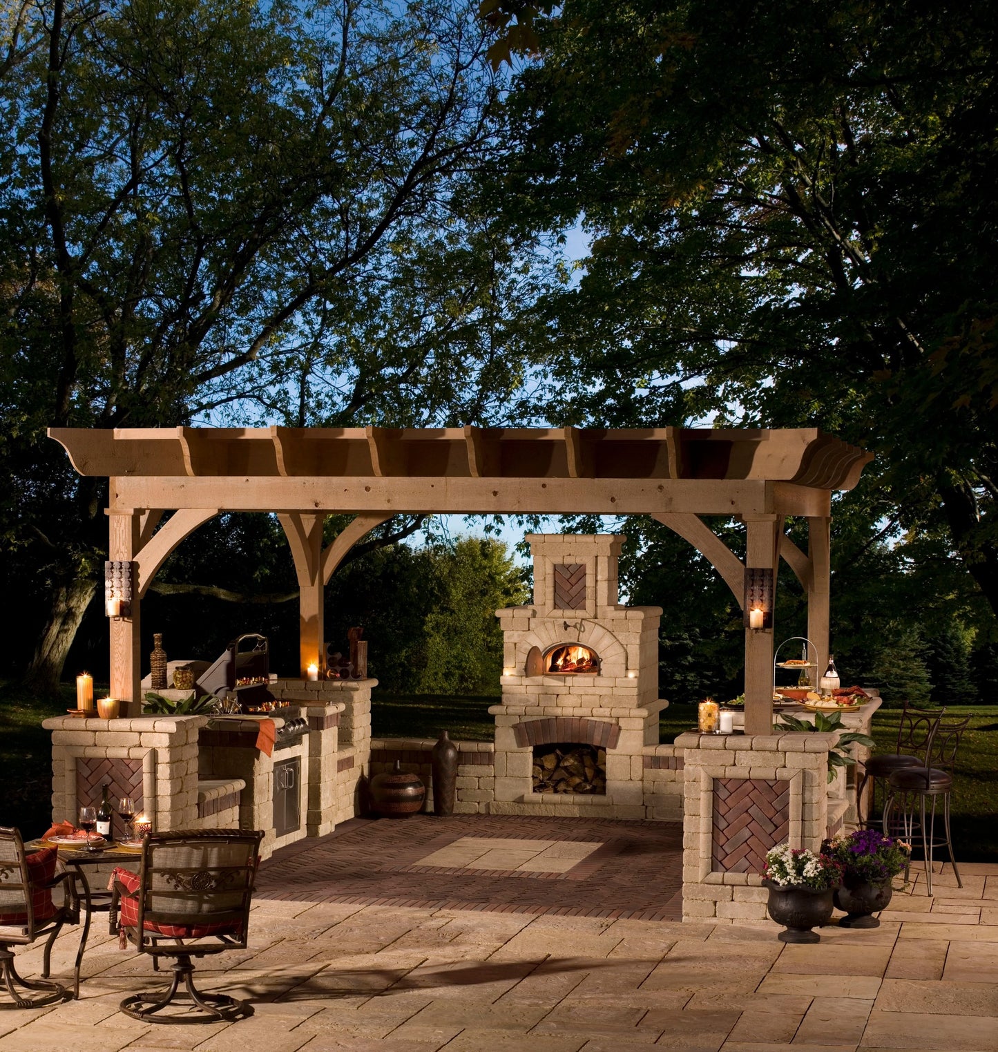 A cozy outdoor kitchen under a wooden pergola features the high-performance CBO 750 DIY Kit from Chicago Brick Oven, a wood-fired pizza oven with a roaring fire, accompanied by a grill station and a stone countertop. The patio is illuminated by lanterns and surrounded by lush trees, creating a warm and inviting evening ambiance.