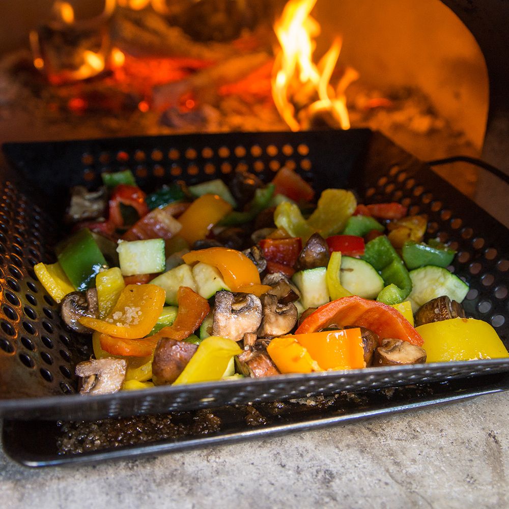 A mix of colorful vegetables, including bell peppers, mushrooms, and zucchini, sizzle in a perforated pan over an open flame. The background reveals a fiery grill with glowing embers from the Chicago Brick Oven CBO 750 Tailgater - Wood Burning Pizza Oven Trailer - RENTAL by Chicago Brick Oven, casting a warm, inviting glow.