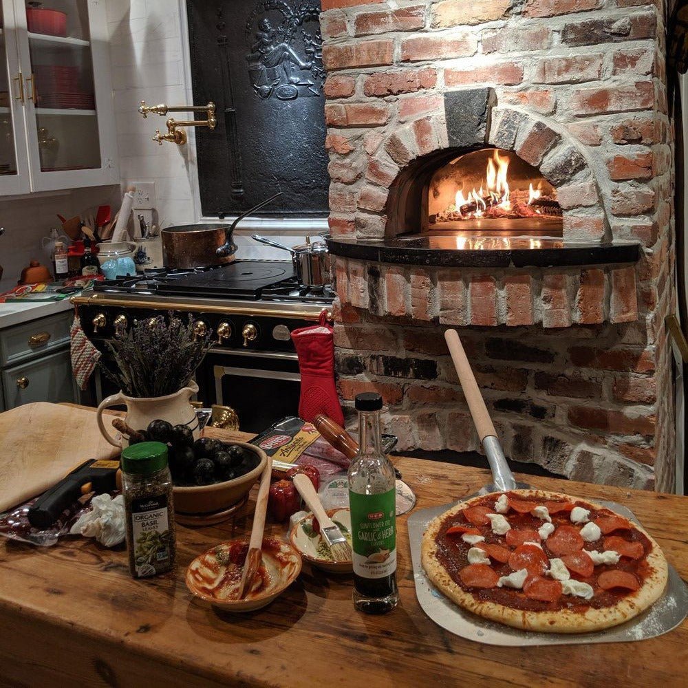 A kitchen with a rustic Chicago Brick Oven CBO 750 DIY Kit, featuring a cozy wood-fired flame inside. A pizza topped with pepperoni and mozzarella rests on a wooden peel on the counter. Surrounding the pizza are ingredients like olives, garlic, and herbs. The CBO 750 DIY Kit promises high-performance cooking in any season, making it perfect for outdoor culinary adventures.