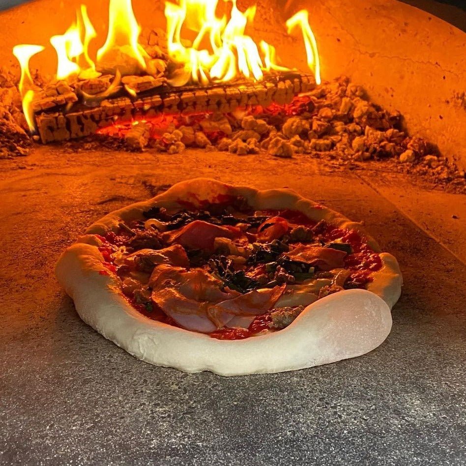Inside a Chicago Brick Oven CBO 750 Tailgater Hybrid Pizza Oven Trailer, a pizza adorned with various toppings is being cooked, with bright flames visible in the background. The edge of the pizza crust appears slightly uneven and puffed. The oven's rustic interior adds to the artisanal feel of the scene.