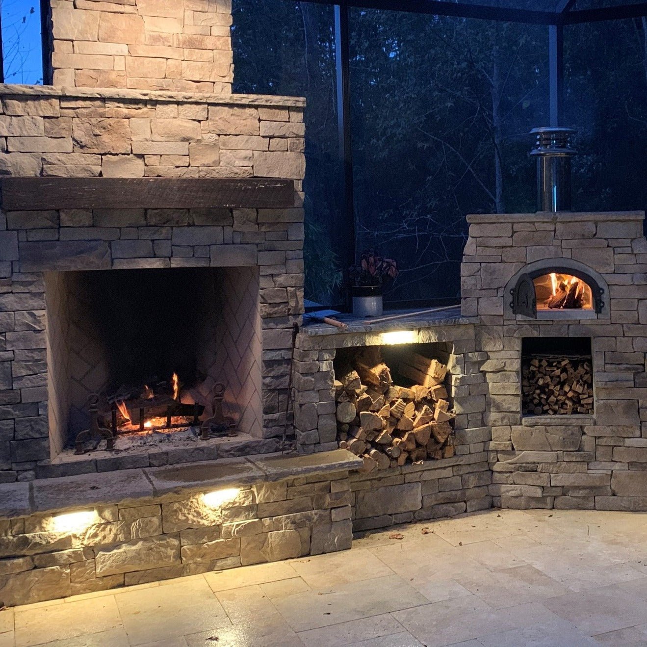A cozy stone outdoor fireplace and oven setup with stacked firewood features the high-performance CBO 750 DIY Kit Wood Fired Pizza Oven by Chicago Brick Oven. This popular bundle, with a 38" x 28" cooking surface, is ready for use beside the burning fireplace. The scene is illuminated by built-in lights, and a dark forest background can be seen through glass panels.