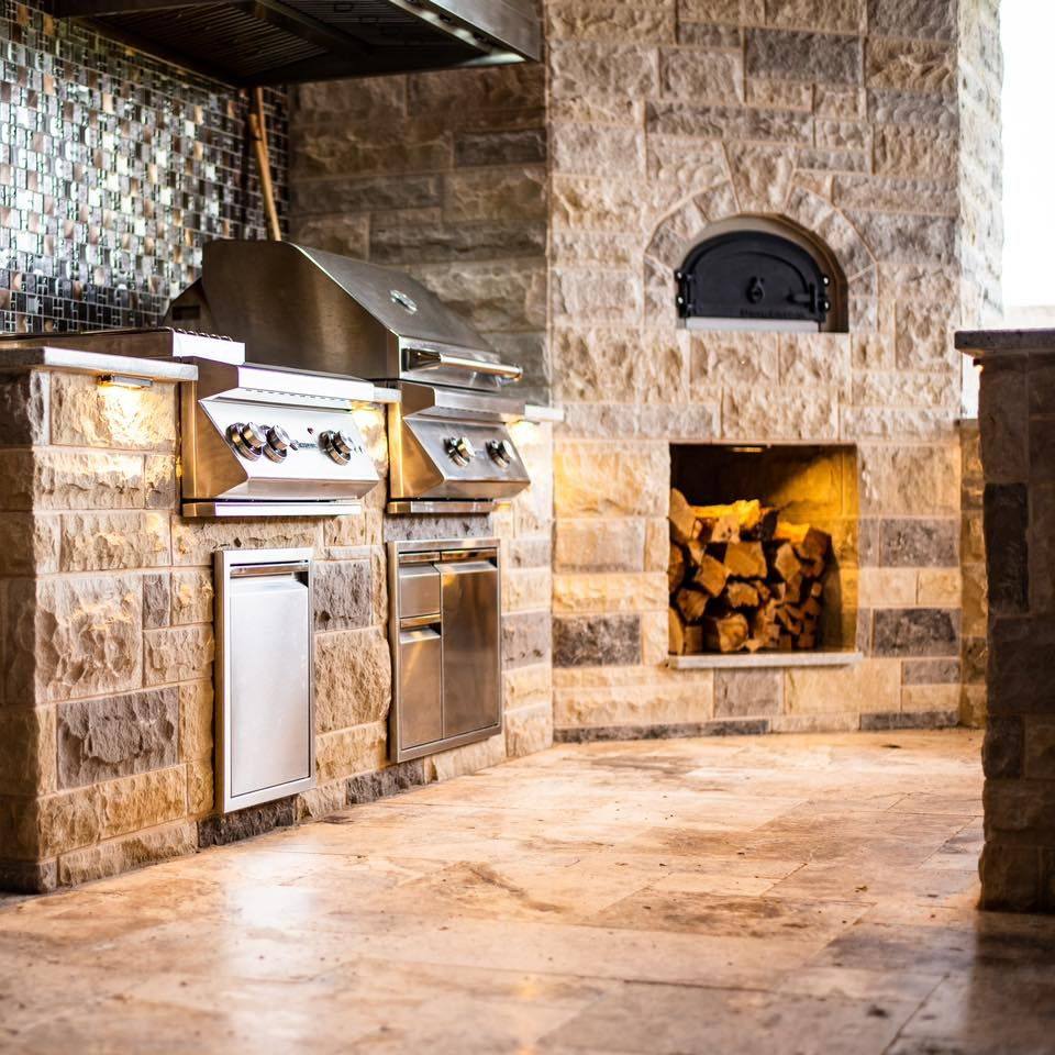 A rustic outdoor kitchen with stone walls features a high-performance stainless steel grill, oven, and storage cabinets. The centerpiece is the Chicago Brick Oven's CBO 750 DIY Kit, our most popular wood fired pizza oven bundle with a 38" x 28" cooking surface. A wood storage nook filled with firewood sits beneath the outdoor pizza oven. The kitchen is accented by mosaic tile backsplash and ambient lighting.