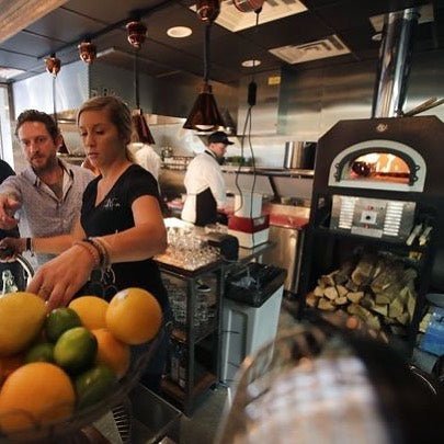 A busy restaurant kitchen with a Chicago Brick Oven CBO 750 Hybrid DIY Kit (Commercial) wood-fired oven. A woman and a man stand near a counter with a bowl of lemons and limes in the foreground. Chefs in the background, using commercial-grade refractory cement ovens from Chicago Brick Oven, are preparing food under copper pendant lights hanging from the ceiling.