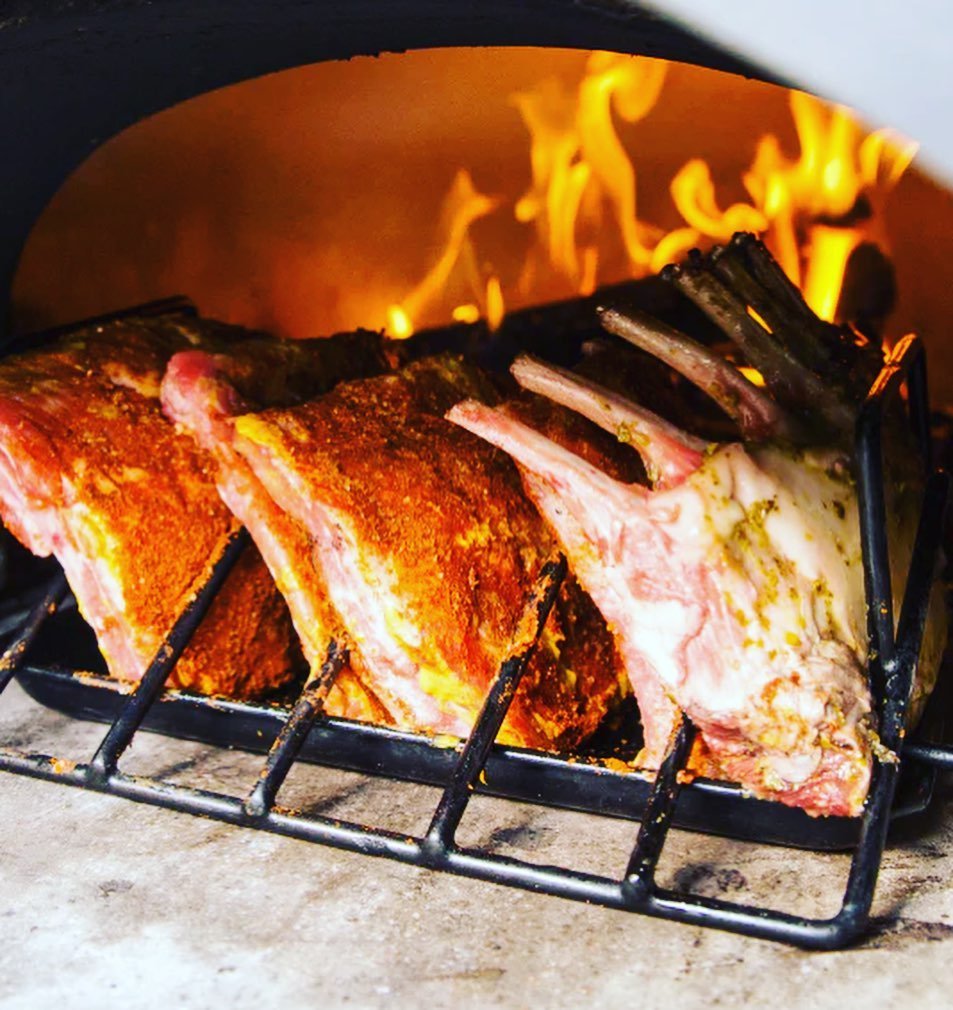 Three seasoned racks of lamb are being roasted on a grill rack inside the Chicago Brick Oven CBO 750 Tailgater, with visible flames in the background. The heat and flames from the hybrid pizza oven suggest the cooking process is well underway, giving the lamb a golden-brown crust.