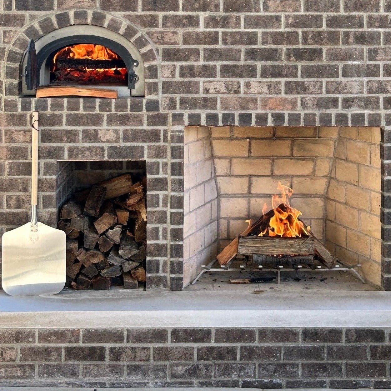 A high-performance brick outdoor kitchen setup featuring the Chicago Brick Oven CBO 750 DIY Kit, our most popular wood-fired pizza oven bundle with a 38" x 28" cooking surface, on the top left with flames visible inside. Below it, a storage space holds firewood. On the right, an open outdoor fireplace has a small fire burning. A pizza peel leans against the structure.