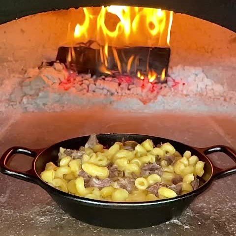 A CBO 750 Tailgater Hybrid Pizza Oven Trailer with a 38" x 28" cooking surface, capable of baking 2-3 ten-inch pizzas at a time, holds a cast iron skillet filled with macaroni and beef inside. The Chicago Brick Oven's interior glows from the heat as flames and embers create a warm, rustic ambiance.