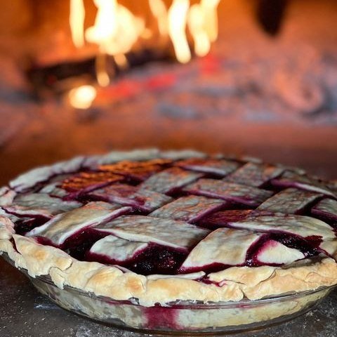 A freshly baked pie with a lattice crust, the filling oozing slightly, sits on a surface in front of the warmly lit, burning fire of the Chicago Brick Oven CBO 750 Tailgater. The pie's golden-brown edge contrasts beautifully with the deep red of its filling, while the wood-fired pizza oven trailer, capable of cooking up to 50-60 pizzas an hour on its expansive 38" x 28" surface, provides a cozy and inviting backdrop.