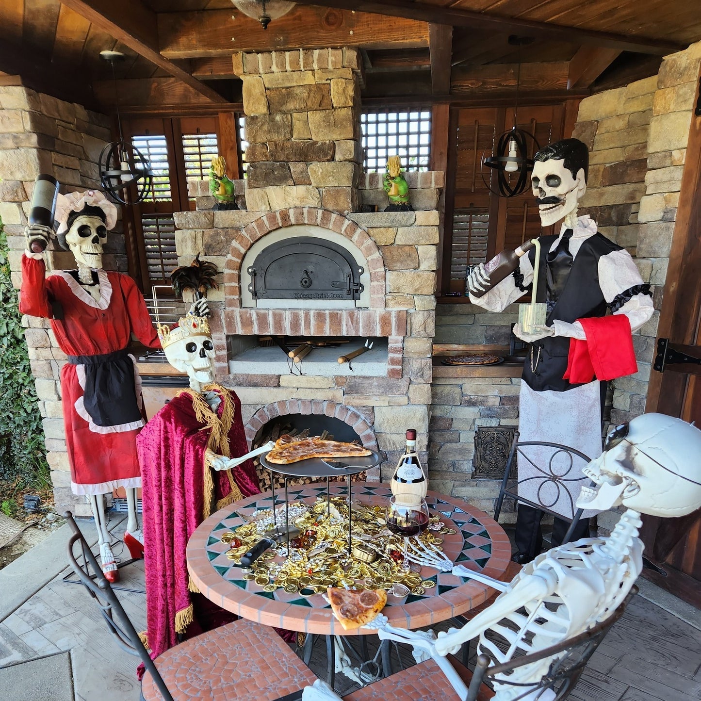 A scene with Halloween skeletons dressed in costumes, sitting around a table with pizza and wine. One skeleton, wearing a red dress, stands with a cleaver, while another in a cape sits. A third skeleton in a waiter uniform holds a tray by the high-performance Chicago Brick Oven CBO 750 DIY Kit, Our Most Popular Bundle Wood Fired Pizza Oven (38" x 28" Cooking Surface). The setting is a cozy, brick-walled outdoor kitchen.