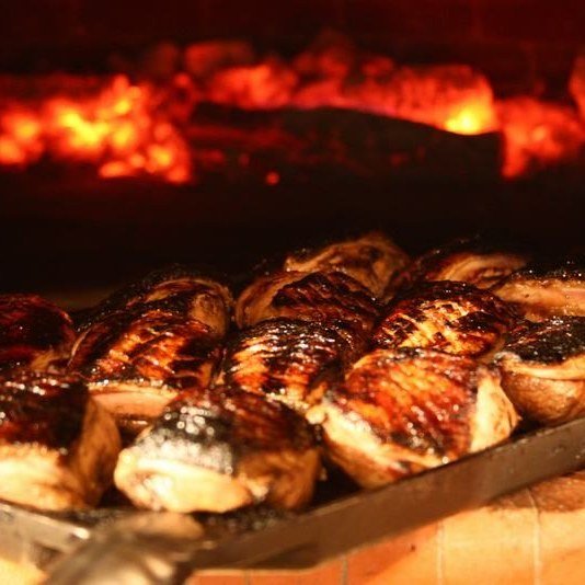 Grilled meats sizzling on a tray over the Chicago Brick Oven CBO 750 Tailgater Hybrid Pizza Oven Trailer, with glowing red coals in the background, creating a warm and rustic atmosphere. The meat is charred and evenly cooked, highlighting the smoky flavors from the open flames of this versatile 38" x 28" hybrid oven that can handle both liquid propane and wood fuels.