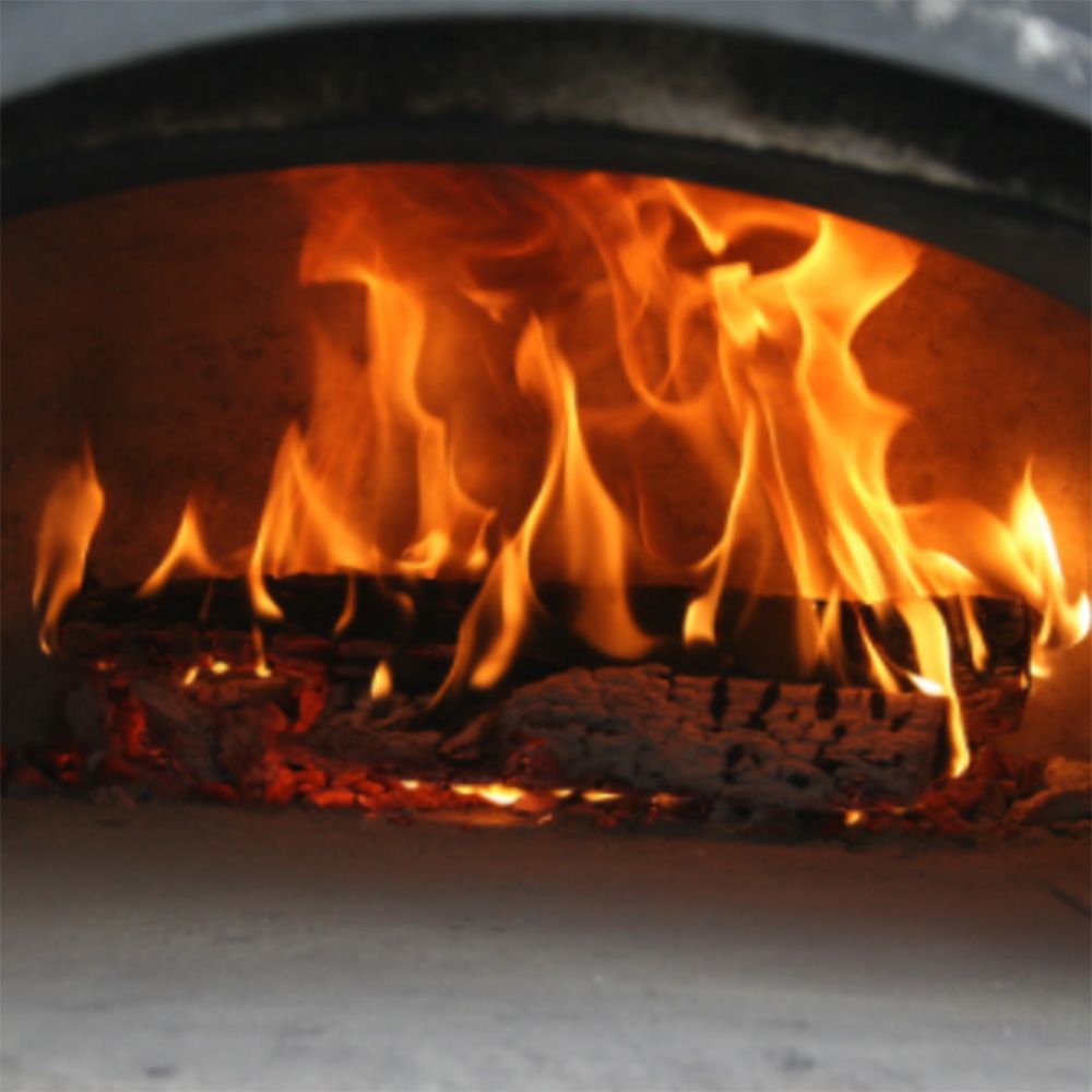 A close-up view of the Chicago Brick Oven CBO 750 Tailgater, with flames dancing on burning logs and casting a warm, orange glow inside this high-performance wood-fired pizza oven trailer. The charred wood and glowing embers enhance the rustic and cozy atmosphere, perfectly complementing its ability to cook 2-3 ten-inch pizzas at a time or up to 50-60 pizzas an hour on its 38" x 28" cooking surface.