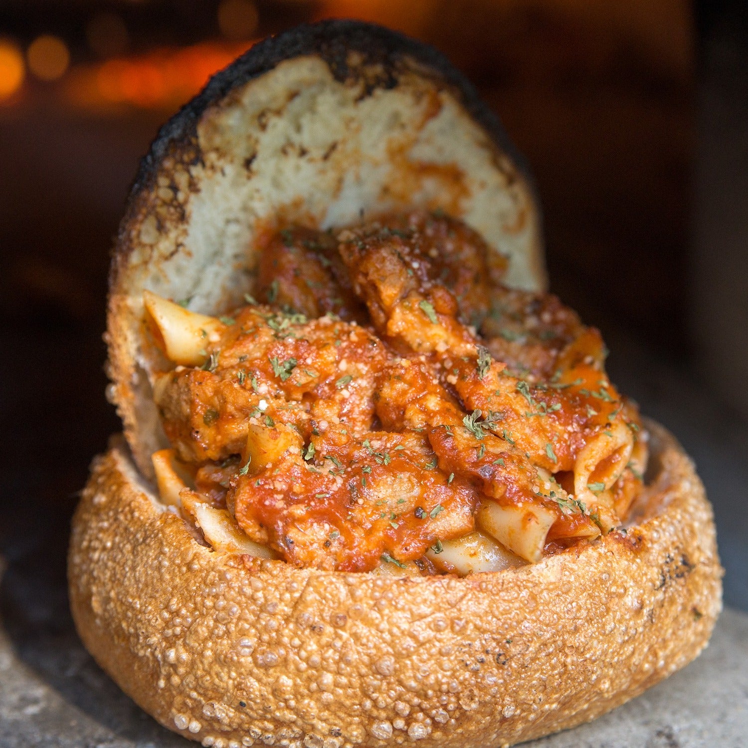 Inside a perfectly baked bread bowl from the Chicago Brick Oven's CBO 500 DIY Kit, hearty pasta is topped with rich tomato meat sauce and garnished with fresh herbs. Baked in a wood-burning oven to crispy, browned perfection, the partially open bread bowl reveals the delicious pasta within. The warm ambiance of a cozy kitchen setting completes the inviting scene.