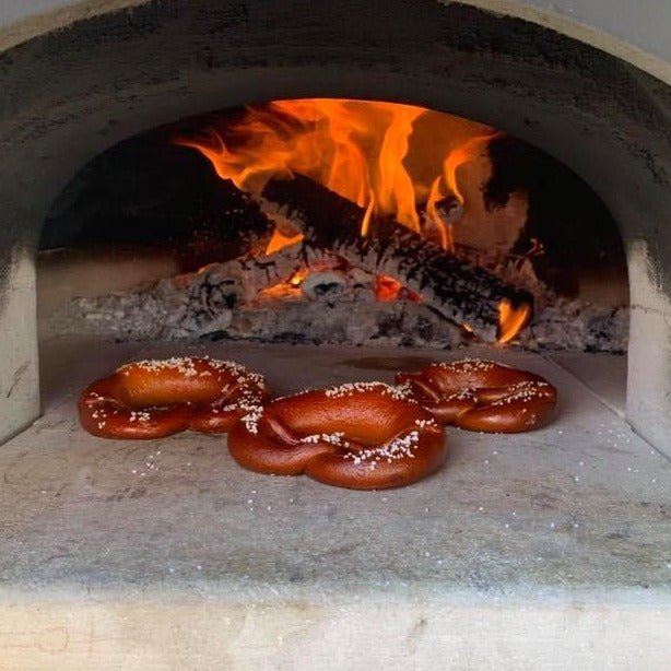 Three pretzels are baking on the stone floor of a Chicago Brick Oven CBO 500 DIY Kit, their golden-brown surfaces sprinkled with coarse salt. A log burns in the background of this wood-fired pizza oven, featuring bright flames and hot embers, perfectly capturing the rustic charm of outdoor cooking. The oven offers flexibility and affordability, boasting a 27" x 22" cooking surface ideal for such delicious creations.
