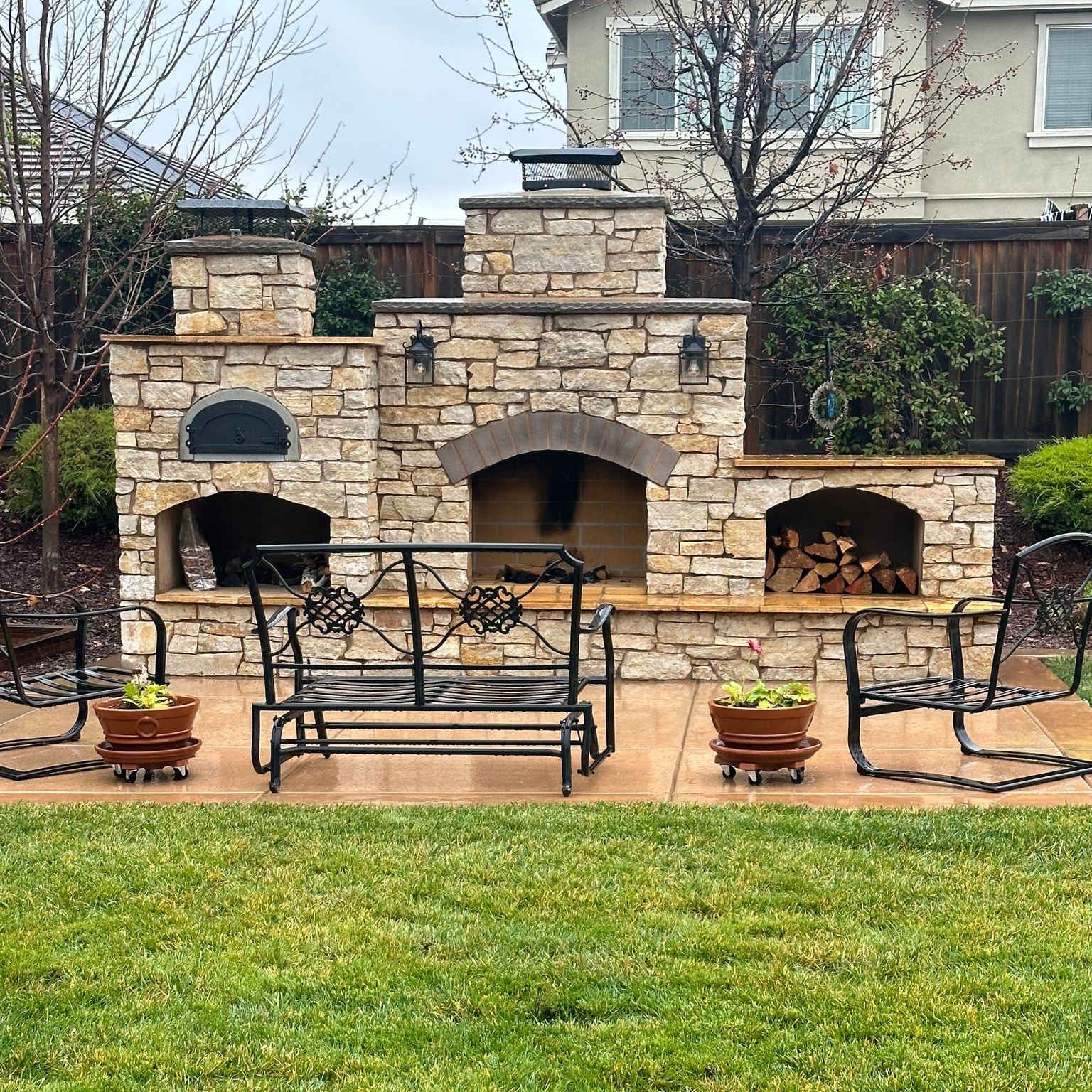 An outdoor stone fireplace and oven setup in a backyard features a Chicago Brick Oven's CBO 500 DIY Kit, boasting a wood-fired pizza oven with a 27" x 22" cooking surface. The setup includes two covered compartments and log storage. Wrought iron patio furniture consisting of a bench and two chairs graces the patio in front, accompanied by two potted plants. Trees and a fence form the backdrop.