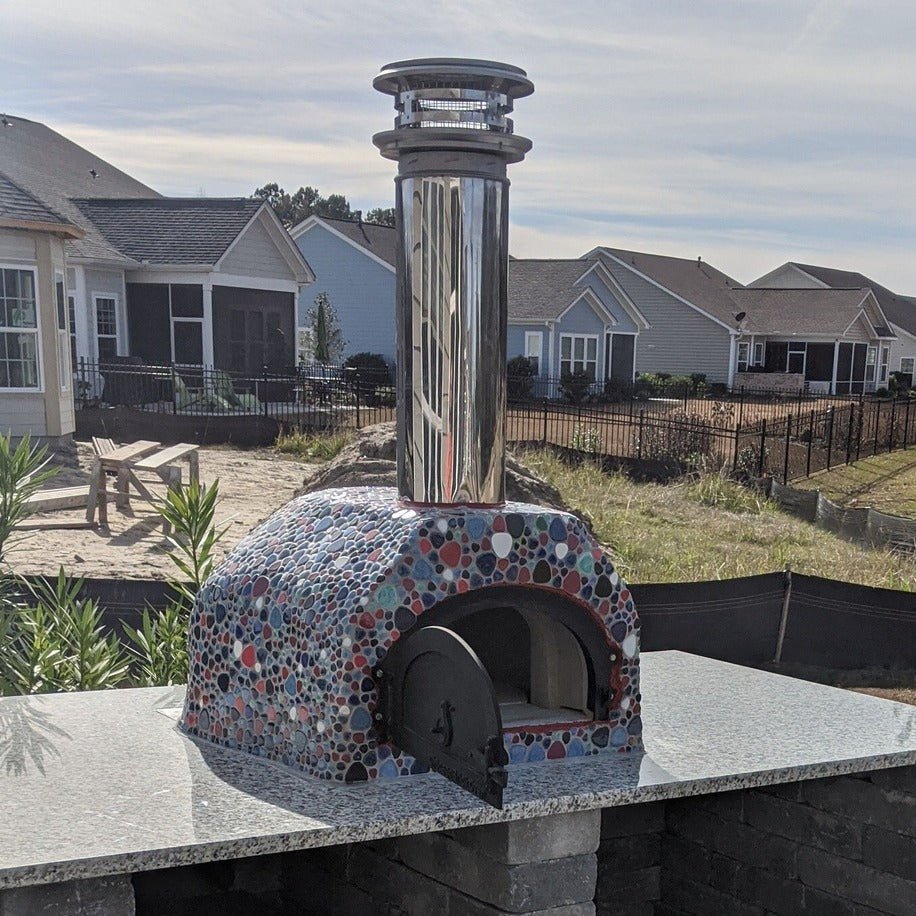 A vibrant mosaic CBO 500 DIY wood-burning pizza oven from Chicago Brick Oven, featuring a stainless steel chimney, sits elegantly on a granite countertop in a residential neighborhood. The oven, with its stone enclosure, is surrounded by houses and fenced yards under clear weather skies.
