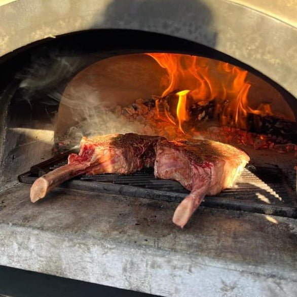 Two large tomahawk ribeye steaks are being grilled inside a CBO 750 Tailgater Wood Fired Pizza Oven Trailer by Chicago Brick Oven. The oven has a visible fire at the back, with flames and burning wood creating a rustic cooking atmosphere. The steaks have a slight sear, indicating they are in the grilling process.