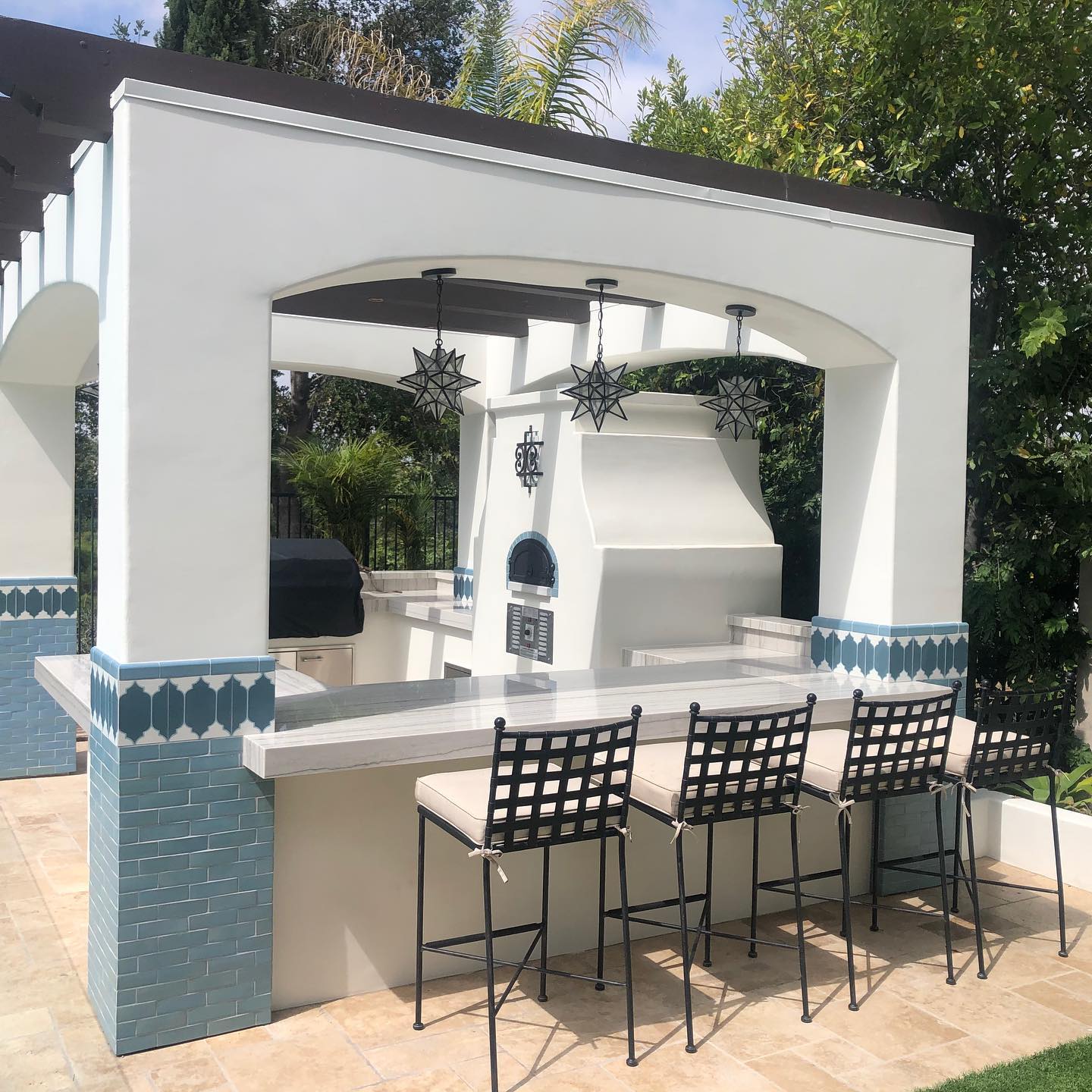 Outdoor kitchen featuring the CBO 750 Hybrid DIY Kit (Commercial) from Chicago Brick Oven, complete with dual-fuel (gas and wood) capabilities. It includes stainless steel countertops and three bar stools, accented by blue tiles and star-shaped pendant lights. The setup is situated on a stone-tiled patio surrounded by lush greenery, ensuring durability with its commercial grade refractory cement construction.