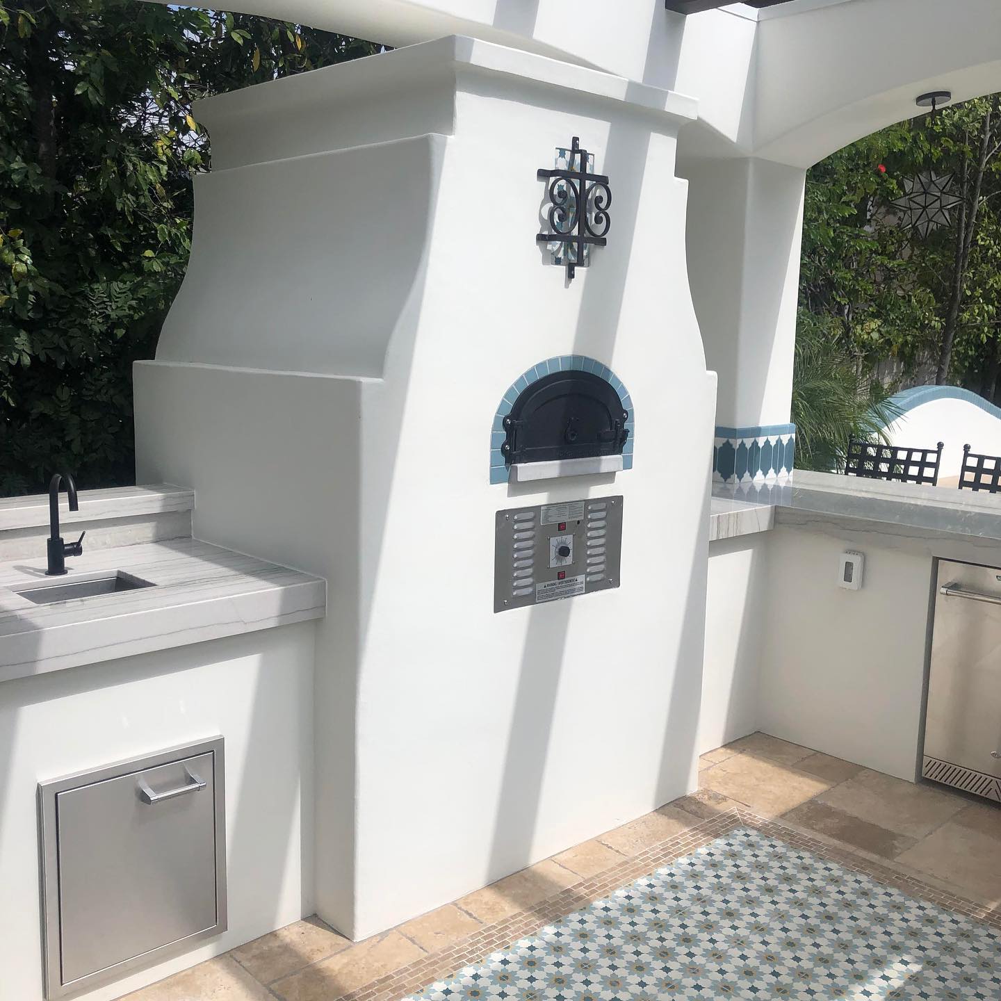 Outdoor kitchen area featuring a Chicago Brick Oven CBO 750 Hybrid DIY Kit with a white stucco finish and a black metal ornament above it, adjacent countertops with a built-in grill, sink, and storage compartments. The space is decorated with tiles and surrounded by lush greenery. The dual-fuel oven, capable of using both gas and wood, is crafted from commercial-grade refractory cement for durability.