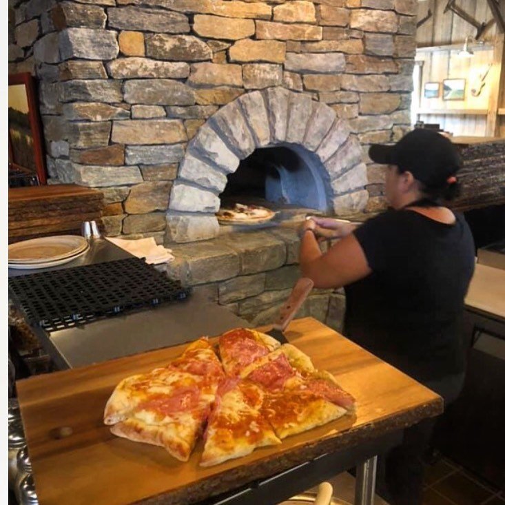 A person wearing a black cap and shirt uses a wooden pizza peel to place or remove a pizza from an outdoor Chicago Brick Oven CBO 1000 DIY Kit wood-fired oven, which boasts an impressive 53" x 39" cooking surface. In the foreground, a wooden board holds a sliced pepperoni pizza, ready to be served. The scene is set in a cozy, rustic pizzeria.