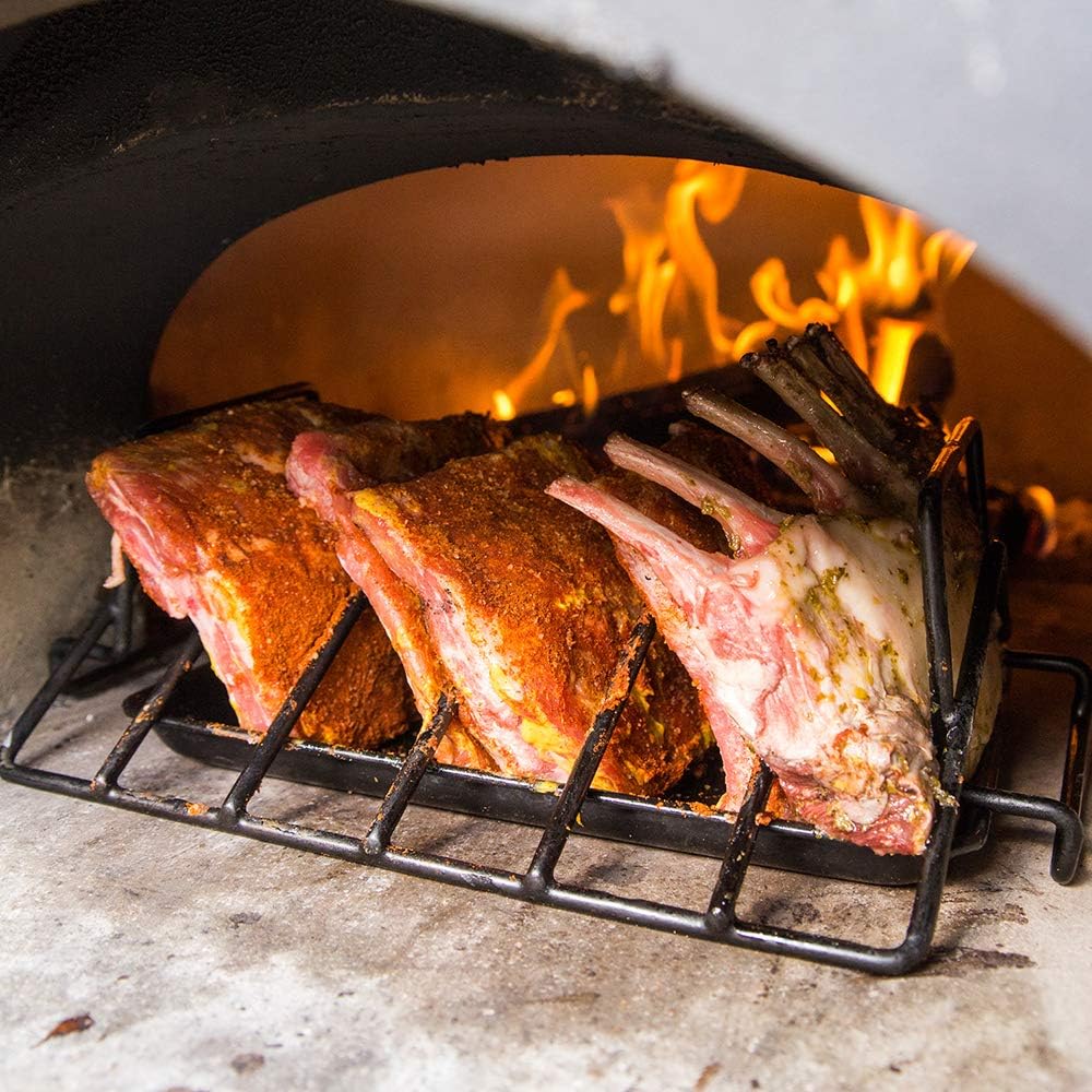 A rack of seasoned lamb ribs is roasting on a grill inside a CBO 1000 DIY Kit | Wood Fired Pizza Oven by Chicago Brick Oven. The flames are visible in the background, creating a warm and rustic atmosphere on the 53" x 39" cooking surface. The meat appears to be well-seasoned and is in the midst of cooking.
