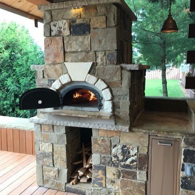 A CBO 750 DIY Kit from Chicago Brick Oven, our most popular bundle, features a high-performance, wood-fired pizza oven with a spacious 38" x 28" cooking surface. This outdoor masterpiece has a sleek black metal door and is beautifully set in a rustic stone structure. Firewood is conveniently stored in the lower compartment of the setup. A hanging light fixture illuminates the area above, with trees providing a serene backdrop.