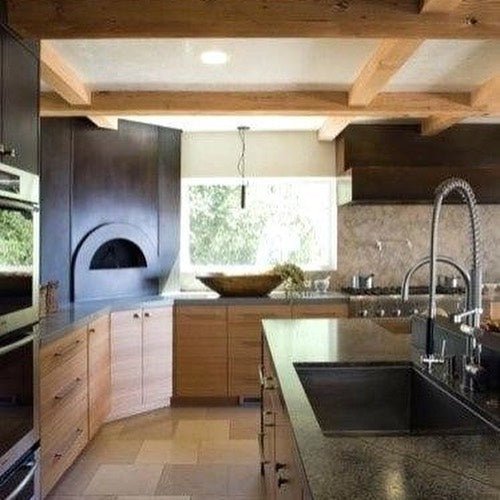 A modern kitchen with wooden cabinets, a built-in high-performance oven, a sleek black countertop, an island with a sink and a high-arc faucet. The kitchen also features a large window, open shelves, exposed wooden beams on the ceiling, and even space for the Chicago Brick Oven CBO 1000 DIY Kit wood-fired pizza oven which boasts a 53" x 39" cooking surface.