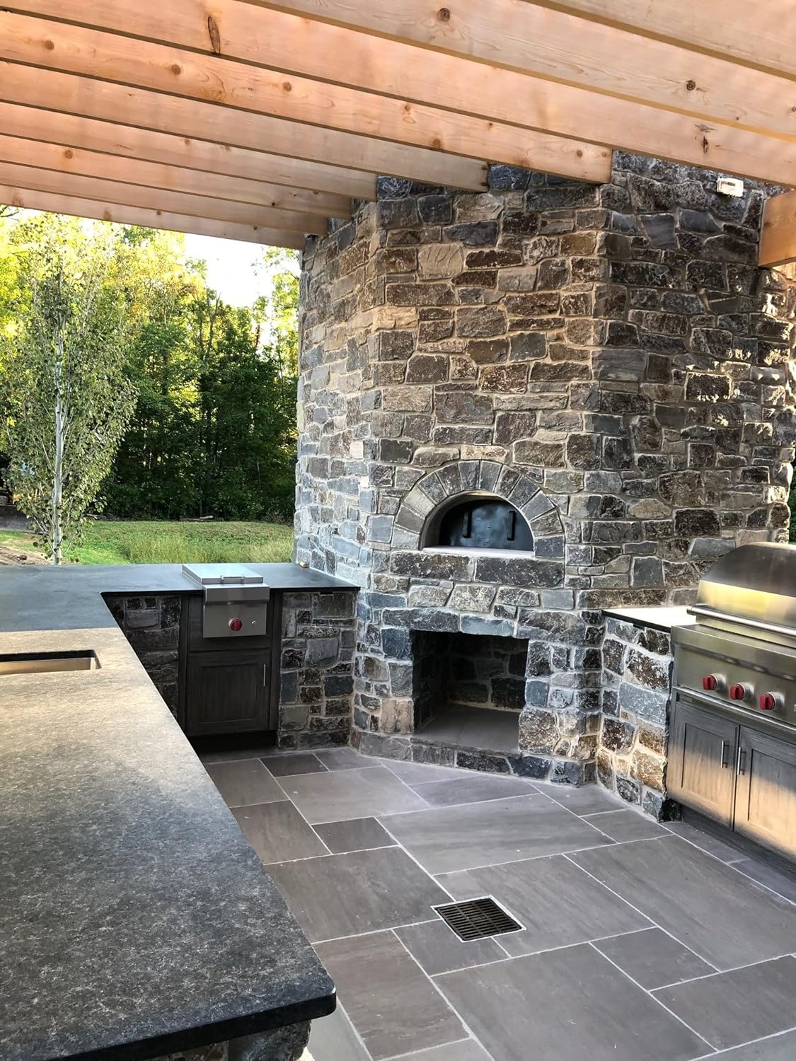 An outdoor kitchen with stone walls featuring the high-performance CBO 1000 DIY Kit by Chicago Brick Oven, boasting a wood-fired pizza oven with a spacious 53" x 39" cooking surface. The space includes countertops, a built-in grill, and cabinetry, all shaded by a wooden pergola overhead. The kitchen overlooks a lush, forested area in the background.