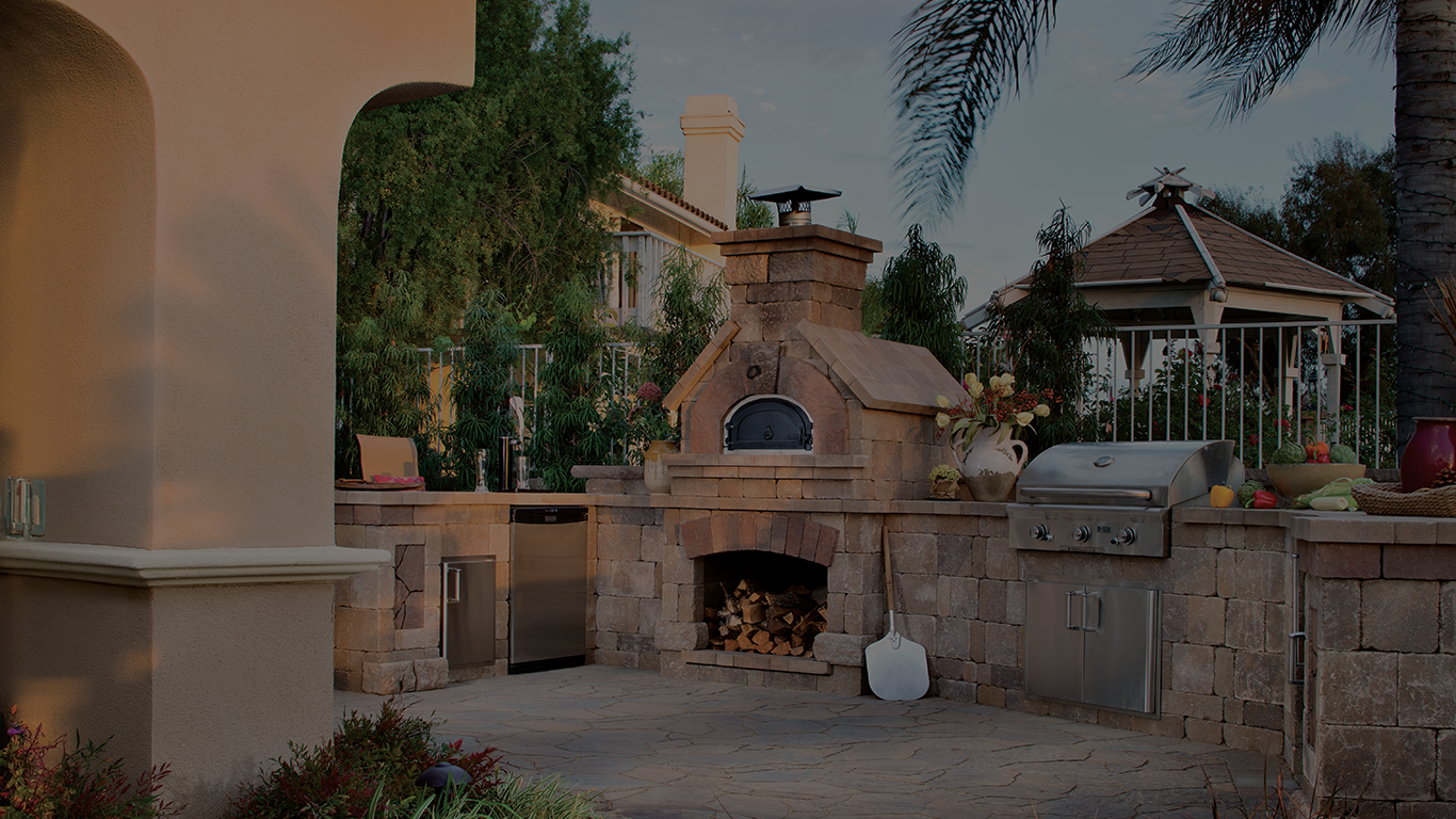 A well-equipped outdoor kitchen with a brick oven, a grill, a prep area, and cabinets. The kitchen sits on a stone patio surrounded by greenery and palm trees. A house with a chimney and a gazebo are visible in the background.