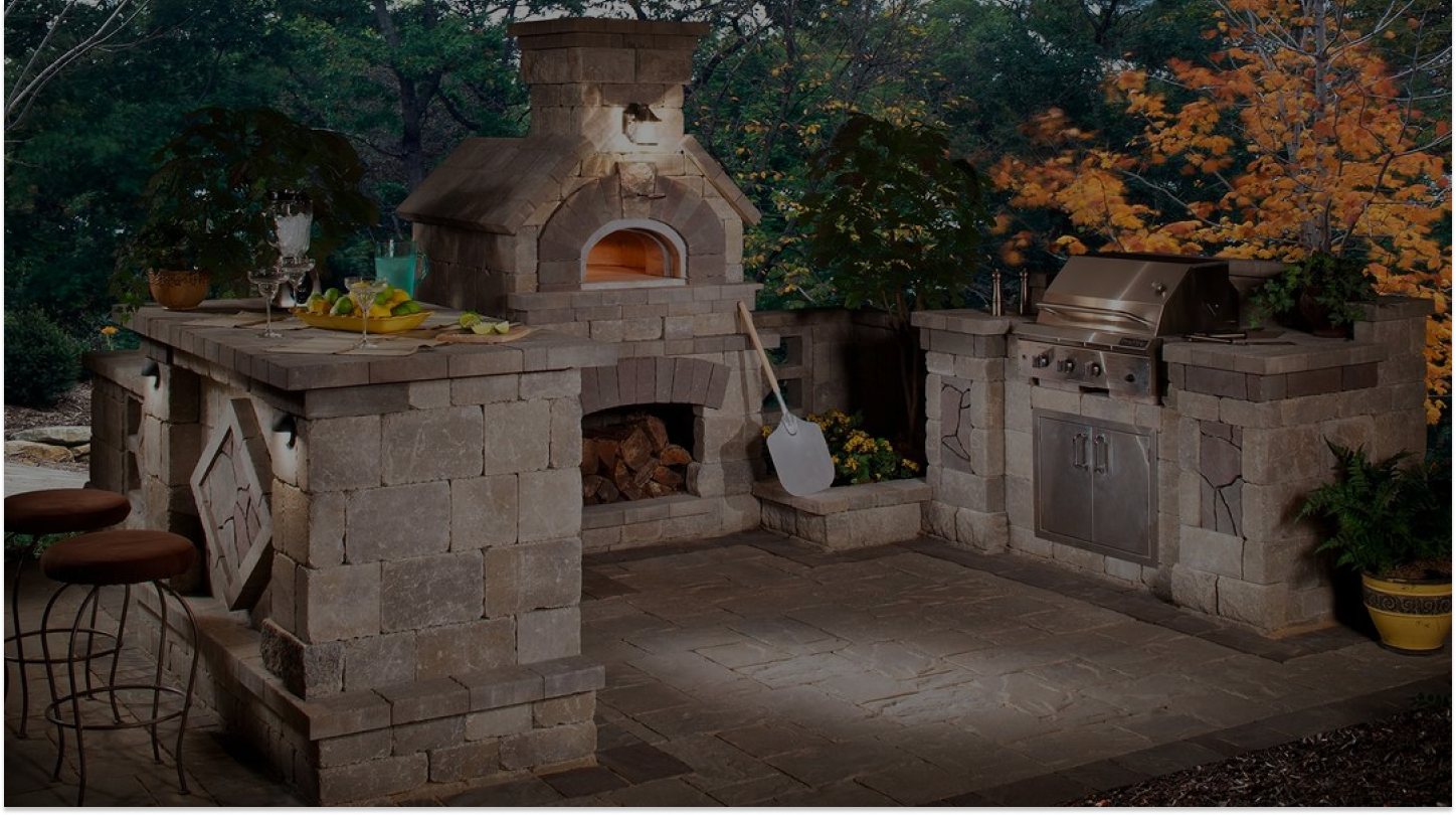 An outdoor kitchen with a stone pizza oven, a grill, and a prep counter with bar stools. There is a pizza peel leaning against the oven, surrounded by trees with green and autumn leaves in the background. The scene is illuminated with soft lighting.