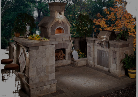 Outdoor kitchen at night with stone counters featuring a wood-fired pizza oven on the left and a built-in grill on the right. The area is softly lit, surrounded by lush greenery and trees with autumn leaves. Wooden stools sit by a counter adorned with plants and corn.