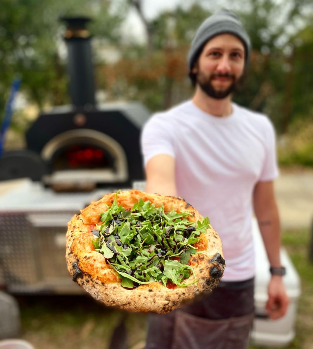 A man with a beard and a gray beanie hat holds a freshly baked pizza topped with greens close to the camera, next to the Chicago Brick Oven CBO 750 Tailgater, an outdoor wood-fired pizza oven trailer. In the background, there is blurred greenery. The man is wearing a white T-shirt and smiling in front of the impressive oven that boasts a 38" x 28" cooking surface, perfect for making 50-60 pizzas an hour or 2-3 - 10" pizzas at a time.