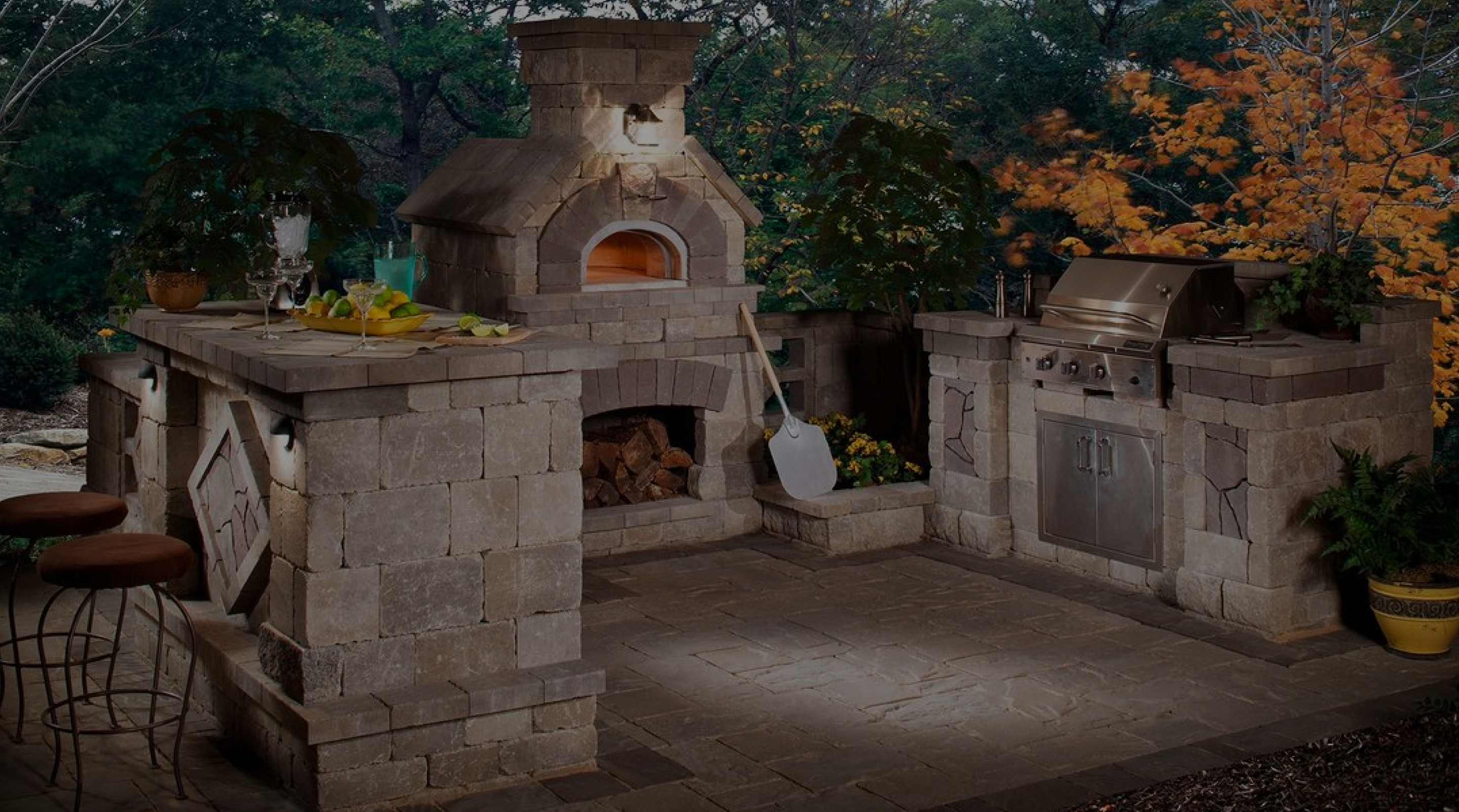 A cozy outdoor kitchen features a stone pizza oven with a paddle, a built-in gas grill, and a bar area with stools. The setup is surrounded by trees with autumn foliage, creating a warm and inviting atmosphere for cooking and dining.