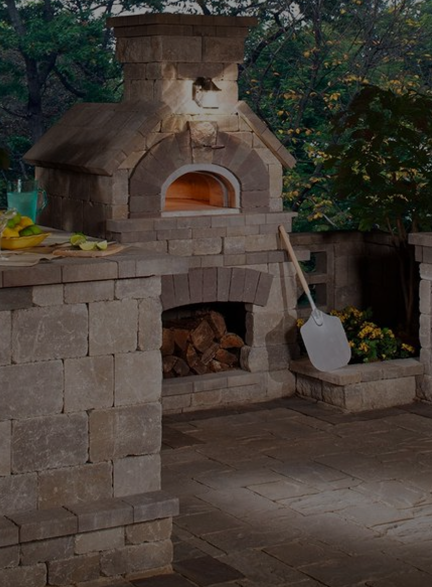 Outdoor stone pizza oven with a wooden door and a chimney, surrounded by a stone counter and walls. A metal pizza peel leans against the counter. Logs are stored in a compartment below the oven. Trees and foliage are visible in the background.