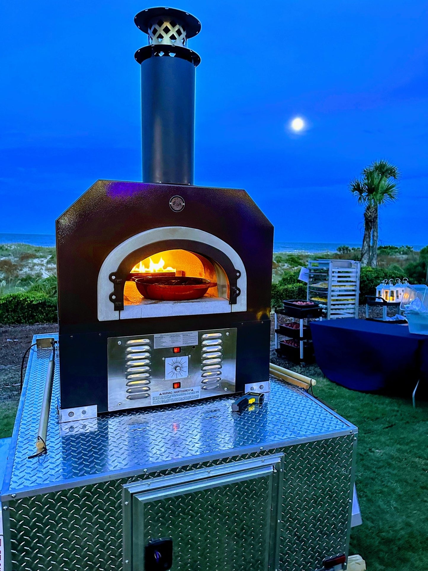 The CBO 750 Tailgater by Chicago Brick Oven, with its black chimney, emits blazing flames while perfectly cooking pizzas under the twilight sky. A full moon shines in the background over a grassy landscape dotted with palm trees and tables set up for an outdoor event. This wood-fired pizza oven trailer features a 38" x 28" cooking surface, allowing you to bake 2-3 ten-inch pizzas at a time and churn out 50-60 pizzas an hour.