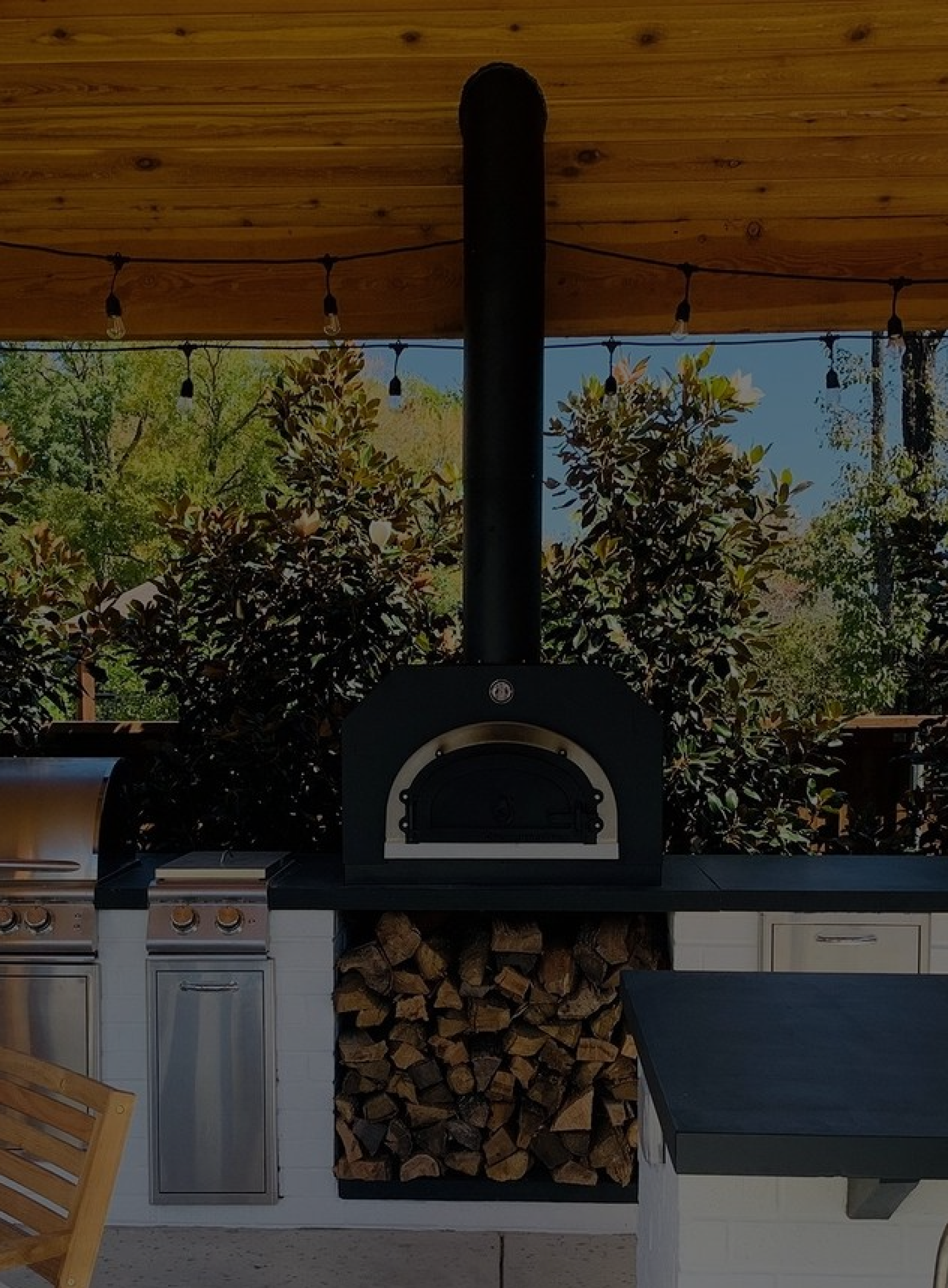 Outdoor kitchen with a wood-fired pizza oven surrounded by trees. The oven is built into a brick countertop with a storage area underneath filled with firewood. A stainless steel gas grill with a side burner is adjacent. String lights hang from the wooden roof above.