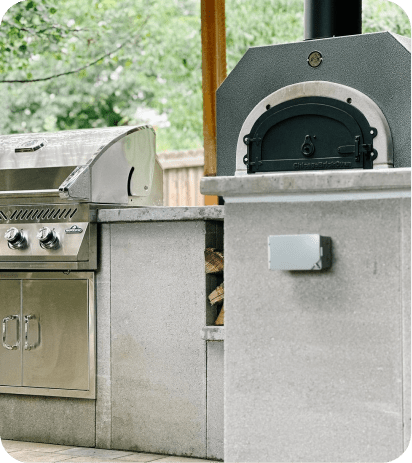 A modern outdoor kitchen featuring a stainless steel grill on the left and the Chicago Brick Oven CBO 500 Countertop wood-fired pizza oven with a 27" x 22" cooking surface crafted from commercial-grade refractory cement on the right. The light-colored stone countertop adds an elegant touch, while a small pile of firewood is neatly stacked underneath the pizza oven. Trees and a wooden fence provide a picturesque backdrop.