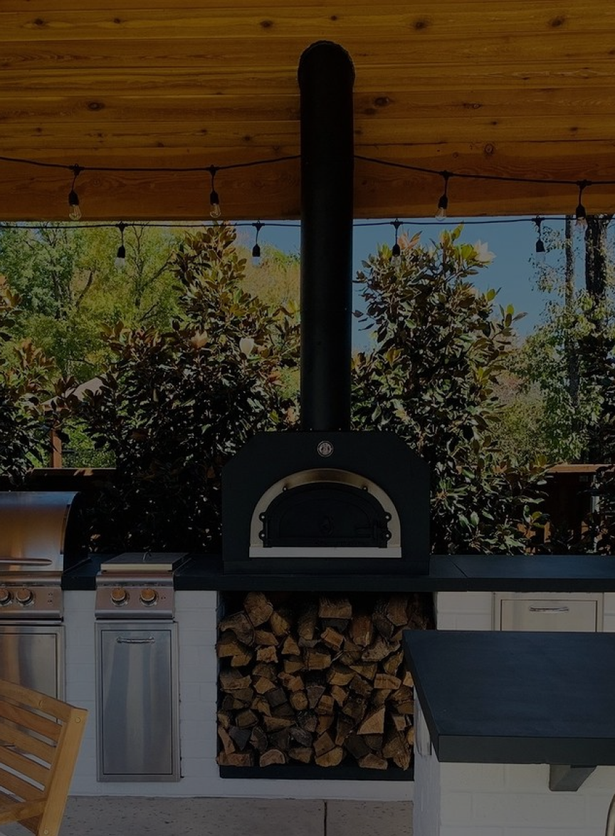 An outdoor kitchen with a pizza oven at the center, a grill on the left, and a countertop on the right. The space includes a wood storage area below the oven and is set against a backdrop of greenery. The kitchen is covered by a wooden roof with string lights hanging.