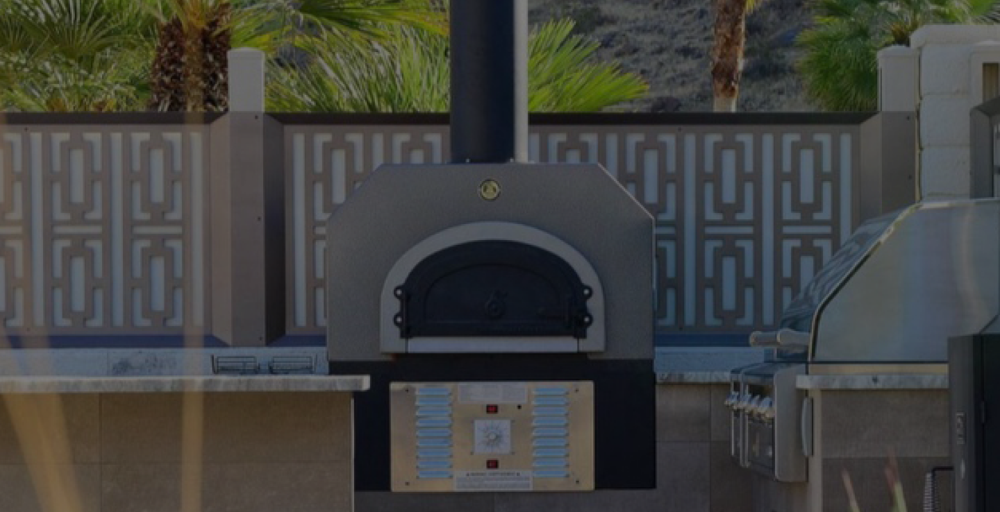 The image shows an outdoor brick and stone pizza oven, complete with a chimney and a decorative metal enclosure. Adjacent to it is a stainless steel grilling station. The background features patterned fencing and palm trees.