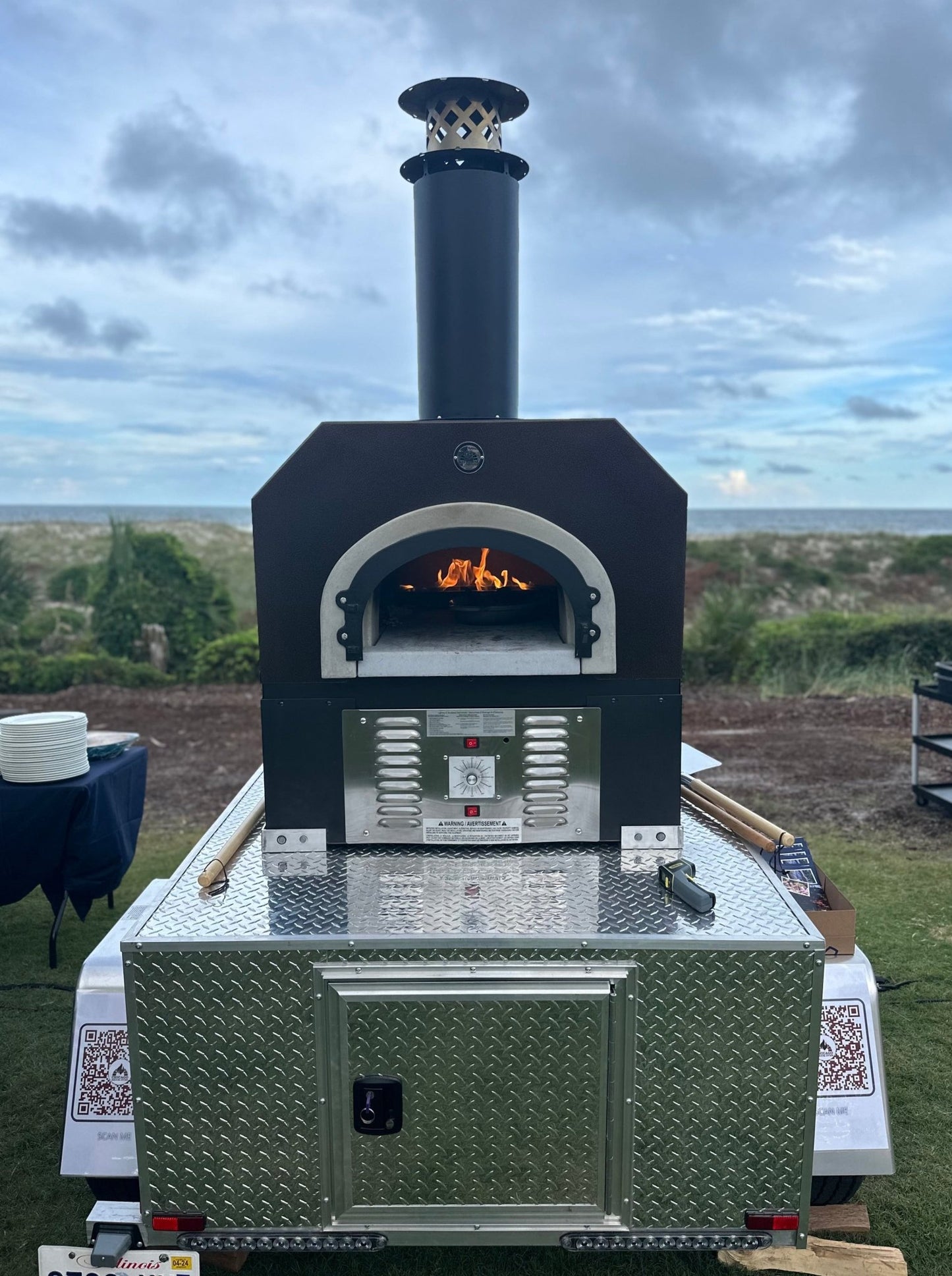 A portable black Chicago Brick Oven CBO 750 Tailgater with a visible flame inside is set up outdoors on a grassy area with a cloudy sky in the background. This high-performance wood-fired pizza oven, which can cook 2-3 ten-inch pizzas at once or up to 50-60 pizzas an hour on its 38" x 28" cooking surface, is mounted on a diamond-plated metal trailer and features various controls and a chimney on top.