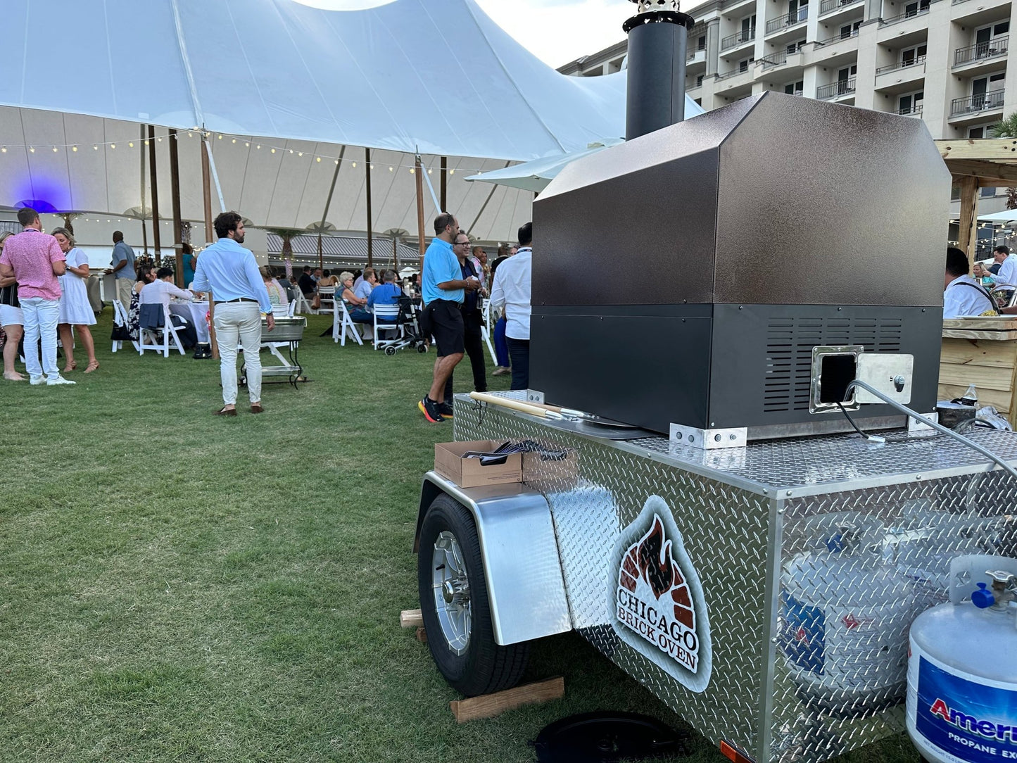An outdoor event is taking place on a lawn with people gathered around tables and standing. A trailer displaying the "Chicago Brick Oven" sign and featuring the CBO 750 Tailgater, a wood-fired pizza oven trailer with a 38" x 28" cooking surface capable of baking 2-3 ten-inch pizzas at a time or up to 50-60 pizzas an hour, is prominently situated in the foreground. A large white tent and building are visible in the background.