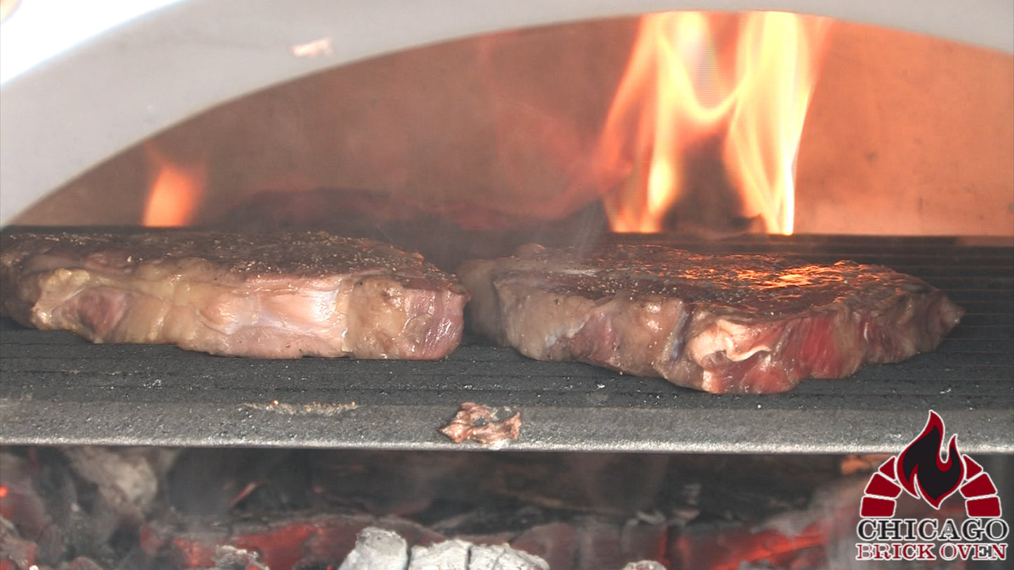 Two thick steaks sizzle on the Tuscan Cast Iron Grill from Chicago Brick Oven, with visible flames dancing in the background. The steaks boast a beautifully seared exterior, suggesting high-heat cooking. In the bottom right corner, a logo reads "Chicago Brick Oven," evoking Steven Raichlen's renowned grilling proficiency.