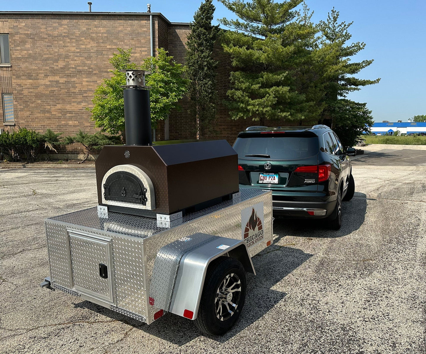 A black vehicle is towing a silver and black CBO 750 Tailgater by Chicago Brick Oven, which is mounted on a small trailer. The wood-burning pizza oven, featuring a chimney and branded with a mountain logo, has a 38" x 28" cooking surface capable of making 2-3 ten-inch pizzas at once or up to fifty to sixty pizzas an hour. The scene is set in an empty parking lot with trees and a building in the background.
