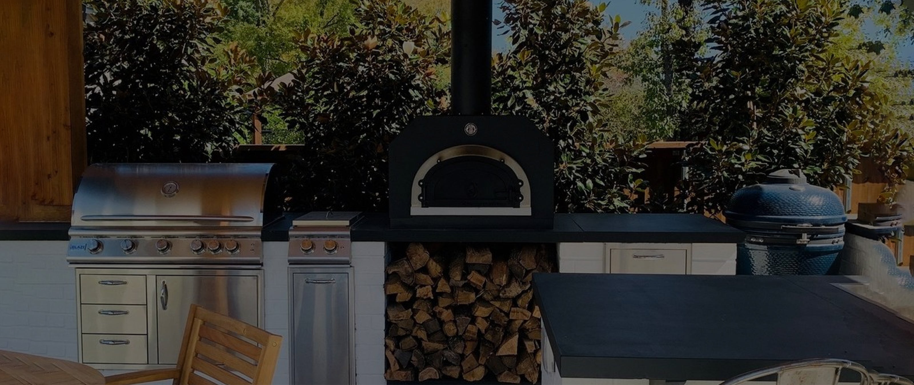 An outdoor kitchen with stainless steel appliances, including a grill, stove, and pizza oven with a wood storage compartment below. A ceramic grill is situated to the right. The background features lush greenery. A wooden dining table is partially visible.