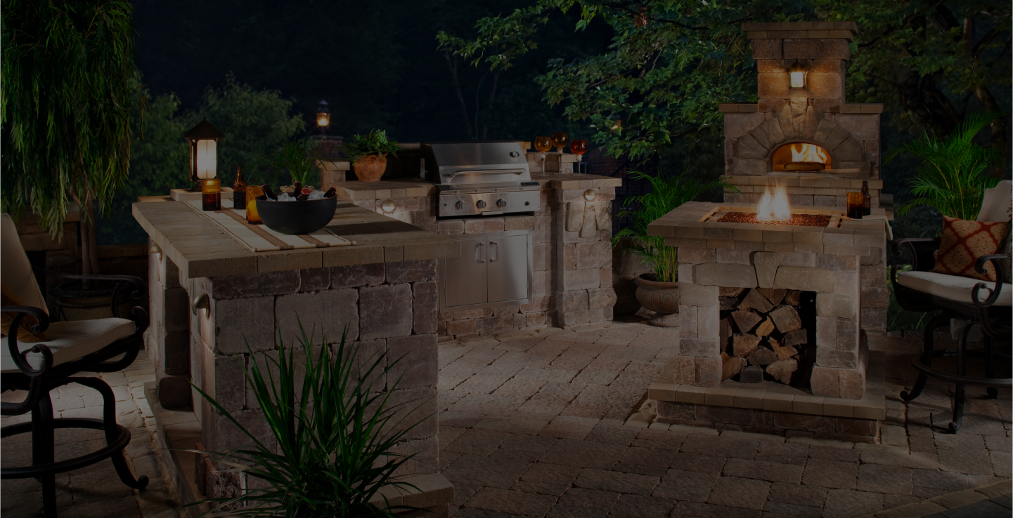 An illuminated outdoor kitchen at night, featuring a stone countertop with a built-in grill, oven, and storage space stocked with logs. The area is surrounded by greenery and has seating with cushioned chairs. Lanterns and potted plants add warmth to the ambiance.